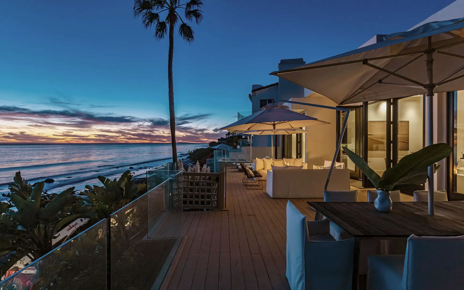 The wood deck at twilight, cream umbrellas over a long outdoor dining table above the beach