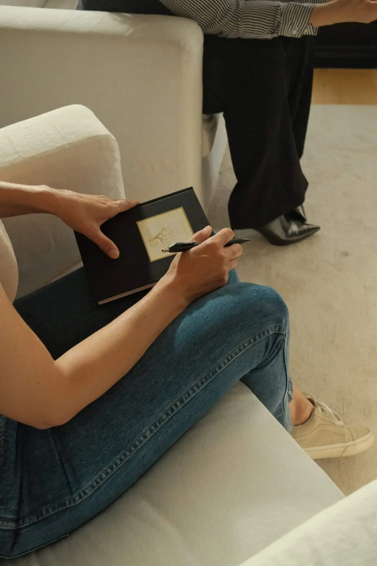 A person seated on a white sofa writing in a notebook, someone standing alongside