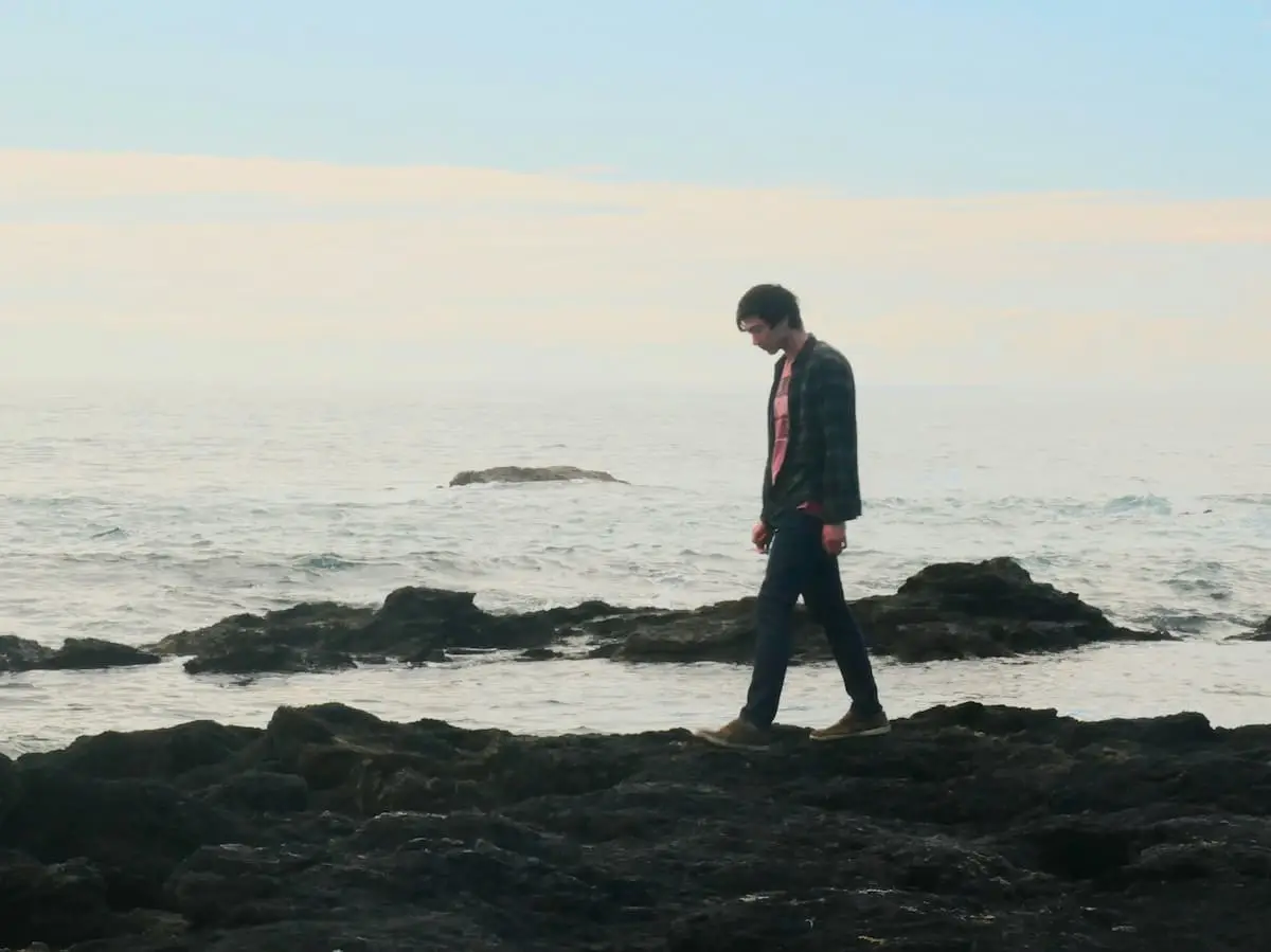 A man walking alone across the rocks at the shoreline, hands in pockets